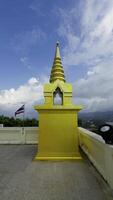 ein Mittel Schuss erfasst ein hell golden Chedi mit ein abgestuft Turm, gelegen auf ein gepflastert Hügel Terrasse. ein thailändisch Flagge Wellen sanft im das Hintergrund, entlang mit üppig Grün Vegetation und ein entfernt Aussicht video