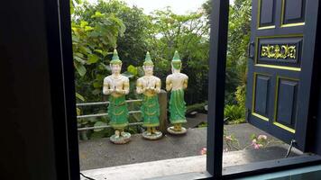 Medium shot from inside a temple looking out through an open dark wooden window at three intricately carved green and gold deity statues standing on a ledge. Lush green foliage and a glimpse of a video