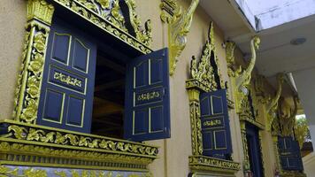 Medium shot of the exterior of a temple showcasing several dark blue window frames adorned with elaborate golden carvings and trim against a light beige wall. Some windows are open, revealing glimpses video