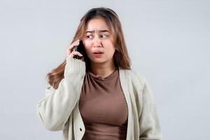 A young woman in a cozy sweater reacts with surprise and confusion while talking on her mobile phone, gesturing with her hand against a neutral background. photo