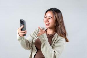 A young woman smiles and points at her smartphone, expressing joy and excitement while engaging with her device in a neutral background. photo