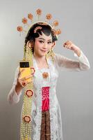 A Indonesian bride in Solo Putri attire, wearing a golden headdress and jasmine garland. She confidently poses with a raised fist and holds a yellow smartphone, blending tradition with modernity. photo