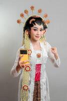 An Indonesian bride in Solo Putri attire, wearing a golden headdress and jasmine garland. She confidently poses with a raised fist and holds a yellow smartphone, blending tradition with modernity. photo