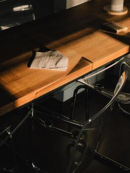 Sunlight illuminates a folded cloth tissue resting on a modern wooden table, with transparent chairs beneath, creating a scene of preparedness and adaptation in a contemporary workspace photo