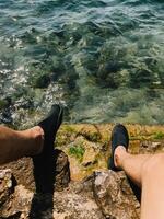 POV shot. Relaxing by sea with feet resting near the water edge. The fresh breeze and warm sun create the perfect coastal escape. Crystal clear sea water reveals rocks below the shimmering surface. photo