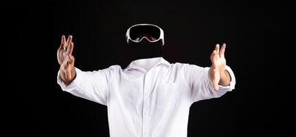 A user in a white shirt is immersed in virtual reality, wearing goggles while gesturing with hands. The background is completely dark, emphasizing the technology and user involvement. photo