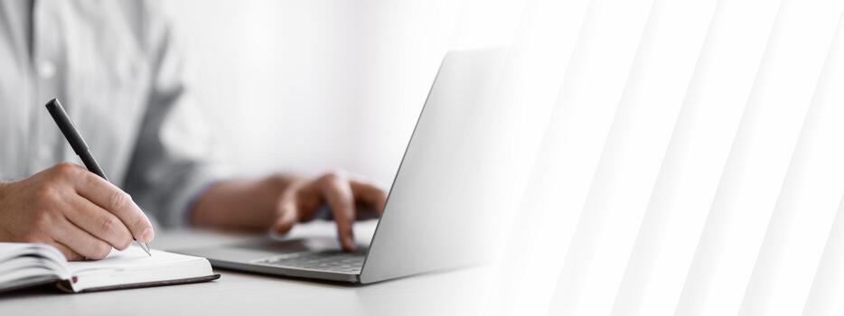 Unrecognizable Man Writing In Notepad And Using Laptop In Office, Taking Notes At Workplace, Using Computer, Noting Information From Internet, Cropped Image With Selective Focus On Hand, Closeup photo