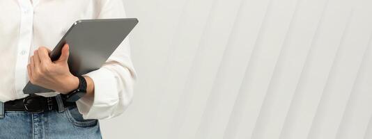 A cheerful millennial european woman with long brown hair and a white blouse holds a tablet and a stylus, looking thoughtfully upwards on a light background, studio. Work, business photo