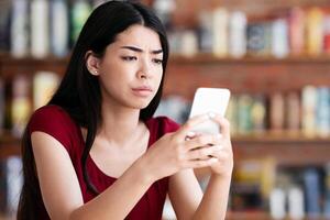 Bad News. Upset Asian Girl Looking At Smartphone Screen In Cafe With Sad Face Expression, Reading Unpleasant Message, Free Space photo