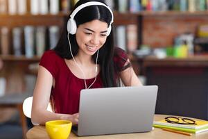 Online Education. Cheerful Korean Female Studying With Laptop And Headphones In Cafe, Watching Webinar Or Online Tutorials, Looking At Computer Screen And Smiling, Enjoying Distance Learning photo