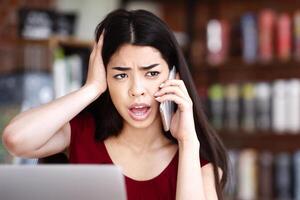 Oh No. Frustrated asian woman having problems with computer, talking on cellphone with customer support while working in remote office in cafe photo