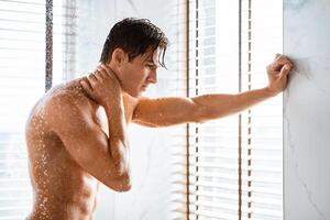 Everyday Routine Concept. Side view of handsome muscular man standing under falling hot water drops in bathroom, leaning on the wall and touching neck, taking refreshing shower and looking down photo