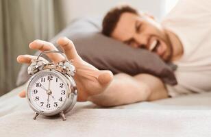 Bad Morning. Angry Man Turning Off Alarm-Clock Waking Up Early Lying In Bed At Home. Selective Focus, Shallow Depth photo