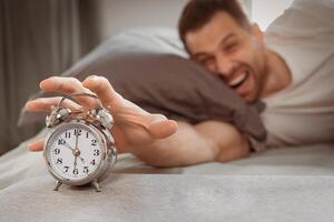 Angry Man Turning Off Alarm-Clock Shouting Waking Up Early In The Morning Lying In Bed At Home, Focus On Clock. Lack Of Sleep, Insomnia And Sleeplessness, Difficult Awakening. Low Light, Shallow Depth photo