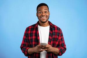Cheerful Black Guy Using Smartphone Smiling To Camera Standing Over Blue Background. Studio Shot photo
