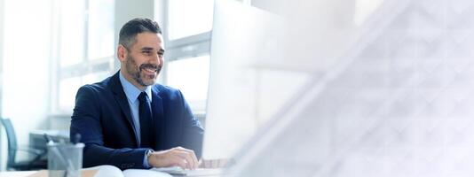 A middle aged Caucasian businessman smiles while working at his desk in a contemporary office. He is focused on his computer, reflecting the essence of a successful and dynamic lifestyle, copy space photo