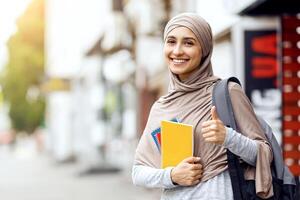 Pretty arab girl with notepads and books showing thumb up, standing on the street, copy space photo