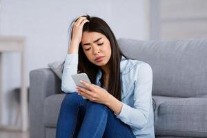 Waiting For His Call. Unhappy Korean Woman Looking At Smartphone Screen At Home While Sitting On Floor In Living Room With Sad Face Expression, Recived Bad Message, Having Depression, Free Space photo