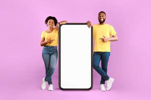 Cheery millennial African American couple pointing fingers at smartphone on violet studio background, mockup for mobile app on white screen. Cellphone website template, cool online store ad photo