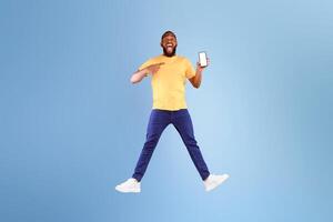Advertisement for mobile application. Excited young African American guy jumping over blue studio background, showing modern smartphone with blank screen, mockup for website or app, full length photo