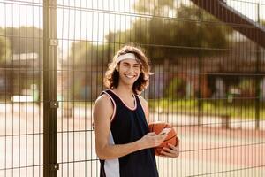 Handsome young sportsman browsing internet on smartphone at outdoor arena, taking break from basketball game, smiling at camera, using mobile device. Professional sports concept photo
