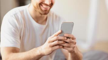 Unrecognizable Smiling Man Using Smartphone Mobile App Sitting In Bedroom At Home, Focus On Phone. Guy Texting On Cellphone With Social Media Application Concept. Cropped, Selective Focus, Panorama photo