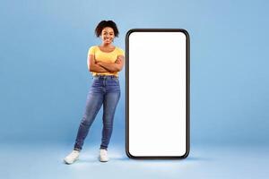 Beautiful African American lady standing near huge smartphone with empty white screen on blue studio background, space for mockup. Newest mobile application, website, advertisement concept photo