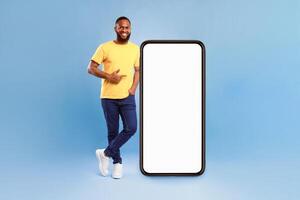 Positive African American guy pointing at big smartphone with mockup for mobile app, recommending cool website over blue studio background. Cellphone screen with space for your advertisement photo