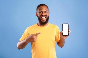 Check out this mobile app. Excited African American man presenting smartphone with blank screen, offering space for website or ad, smiling at camera on blue studio background, mockup photo