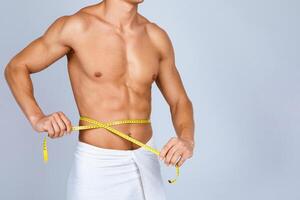 Fitness And Healthy Lifestyle Concept. Closeup of unrecognizable sporty muscular man measuring his waist with yellow tape. Young guy standing wrapped in towel isolated over studio background photo