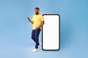 Handsome young African American man using mobile phone while standing near huge smartphone with empty screen over blue studio background. Mockup for website or application, space for advertisement photo
