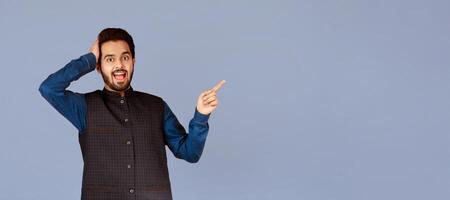 Excited Indian guy holding his head in disbelief and pointing at empty space over violet background photo