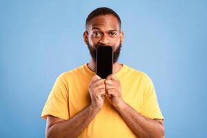 Handsome African American man covering is mouth with blank cellphone with mockup for app or website design over blue studio background. Cool guy showing mobile advertisement photo