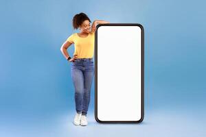 Joyful African American woman in casual wear leaning on huge cellphone with empty white screen on blue studio background, mockup for mobile app, website, your ad design. Smartphone template photo