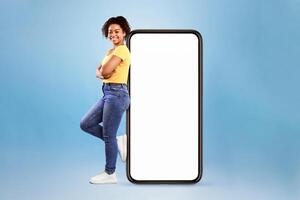 Full length of joyful African American woman leaning on big cellphone with empty space for mobile app on white screen, blue studio background. Smartphone display mockup, website or ad template photo
