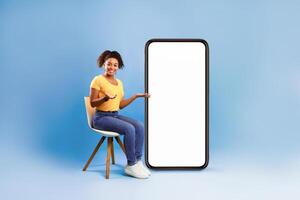 Full length of pretty African American lady sitting on chair next to big blank smartphone with mockup for mobile app or website design, blue studio background. Space for your online advertisement photo