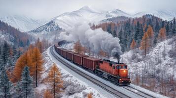A freight train transports cargo containers through a snow covered mountain pass surrounded by autumn foliage. The locomotive emits smoke as it travels along the winding tracks. photo