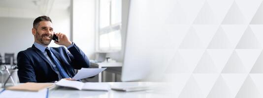 A middle aged businessman wearing a suit smiles while talking on the phone at his office desk. He holds a document, signifying his active engagement in work and success in entrepreneurship, copy space photo