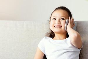 Phone Call. Asian Little Girl Talking On Cellphone Sitting On Sofa At Home. Empty Space For Text photo
