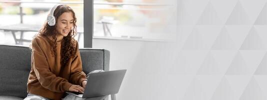 E-Learning. Cheerful woman student using laptop computer, typing on keyboard at home, wearing wireless headphones. Lady on sofa websurfing and learning online. Free space, panorama photo