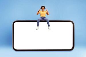 Positive millennial African American woman showing thumb up gesture, sitting on top of huge smartphone over blue studio background, empty space for mobile app on screen. Website or advert mockup photo