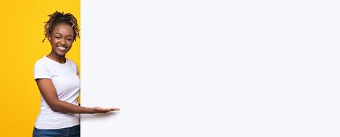 Smiling black woman with white empty board for advertisement over yellow background, panorama photo