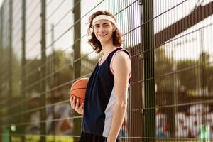 Portrait of positive millennial basketball player in sports uniform posing at outdoor stadium before game, empty space. Young basketballer smiling at camera after training for big competition photo