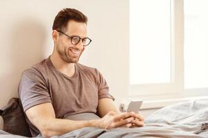 Smiling Man Using Smartphone Mobile App Sitting In Bed At Home On Weekend Morning. Free Space For Text photo