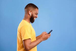 Side view of millennial African American man using smartphone, communicating online in earphones on blue studio background, copy space. Cool guy having virtual meeting on mobile device photo