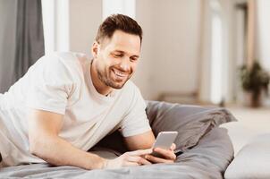Happy Man Using Smartphone Lying In Bed At Home. Mobile Application. Copy Space photo