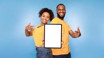 Cheerful African American couple pointing at digital tablet with blank white screen, recommending new application on blue studio background, mockup for design. Website, ad template photo