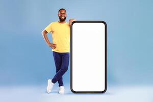 Full length portrait of cheerful African American guy leaning on giant smartphone, demonstrating empty screen on blue studio background, copy space for online advertisement, mockup for mobile app photo