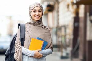 Muslim girl going to university, holding notepads and smiling at camera on the street, educational concept photo