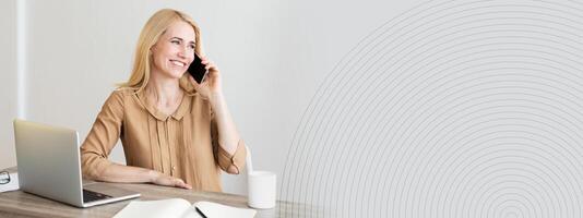 A woman engages in a phone conversation while sitting at a contemporary desk with a laptop and an open notebook. The workspace is well lit, creating a pleasant atmosphere for productivity, copy space photo
